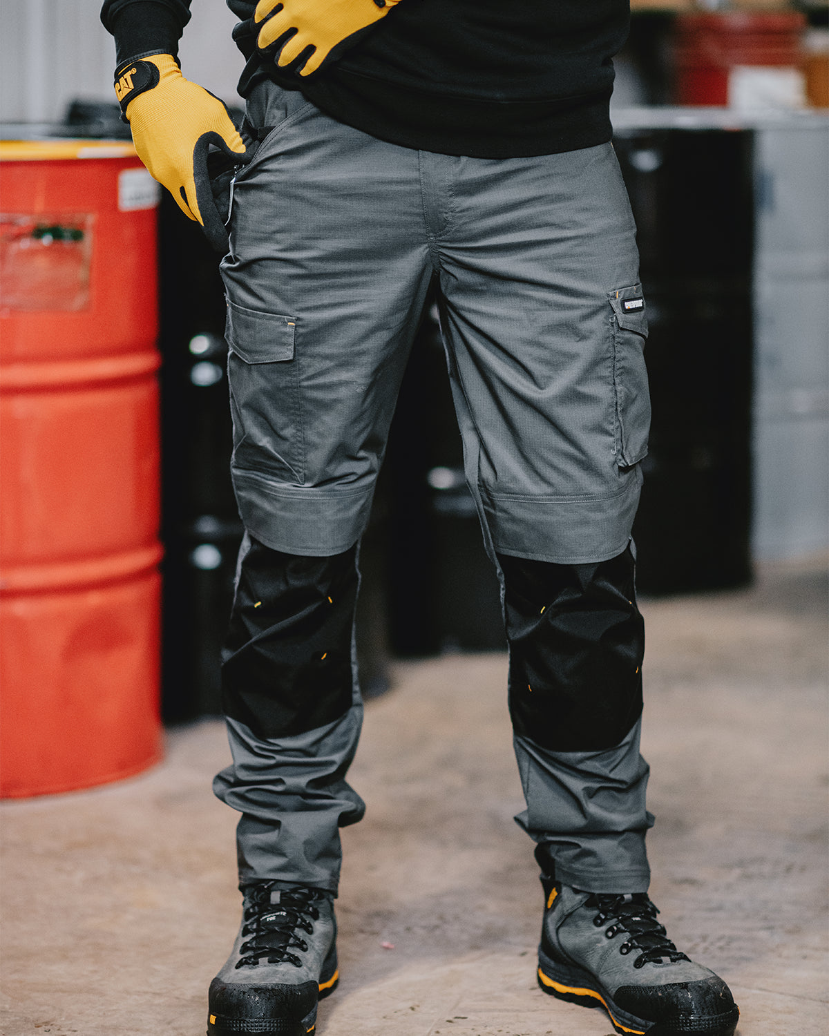 Cat workwear Men's Nexus Work Pant Dark Shadow. Person wearing gray work pants, black boots, and yellow gloves in an industrial setting.